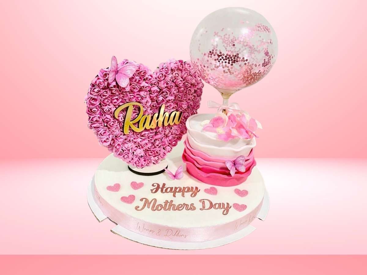 Decorative cake with pink heart-shaped cake topper, 'Happy Mothers Day' text, and balloons on a pink background.